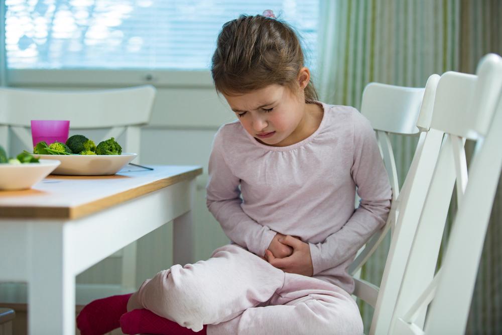 Menina com intoxicação alimentar, sentada à mesa de jantar, reagindo à dor, colocando as mãos na barriga. 