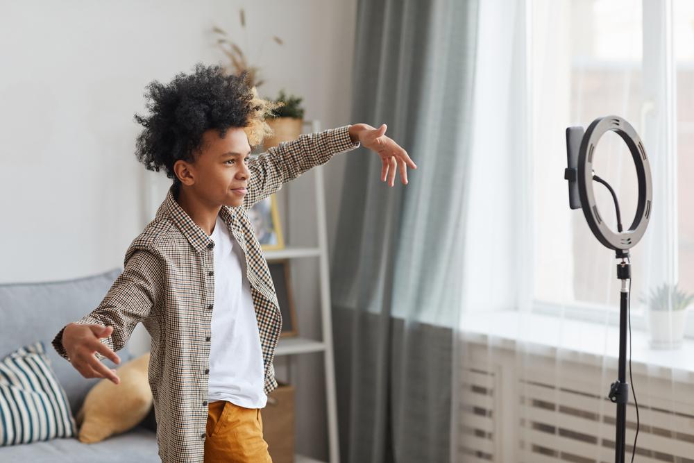 Um menino está fazendo uma pose de braços abertos em frente ao celular, que está apoiado em um tripé com ring light. 