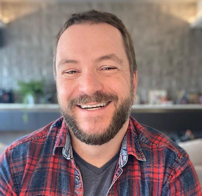 A foto mostra Beto Bigatti sorrindo em ambiente interno e iluminado usando uma camisa xadrez.