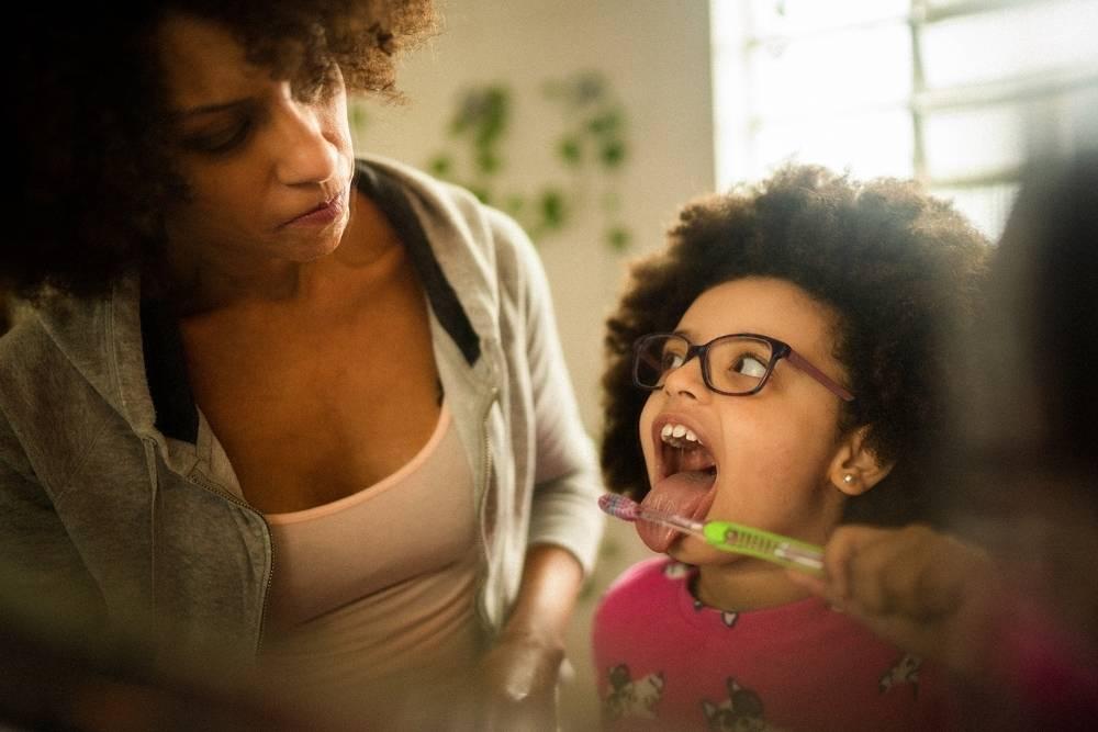 Uma menina está escovando os dentes, parte importante dos hábitos de higiene, enquanto uma mulher adulta a ajuda.