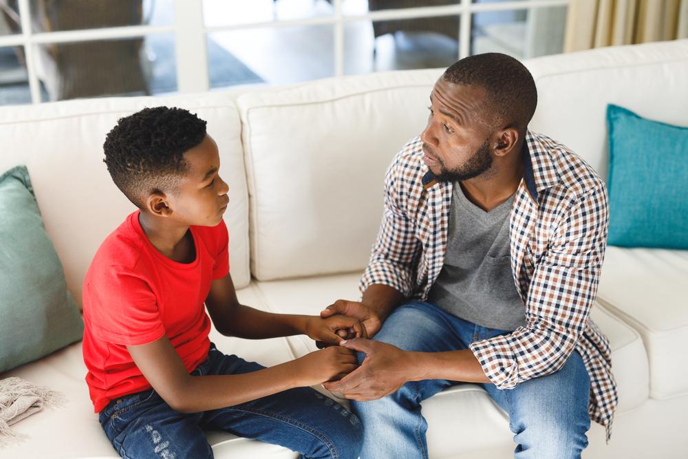 pai e filho sentados no sofá da sala de mãos dadas se olhando e conversando com expressões sérias