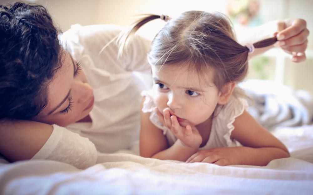 Mãe e filha deitadas na cama, onde a mãe alisa os cabelos da criança e a mesma aparenta estar reflexiva