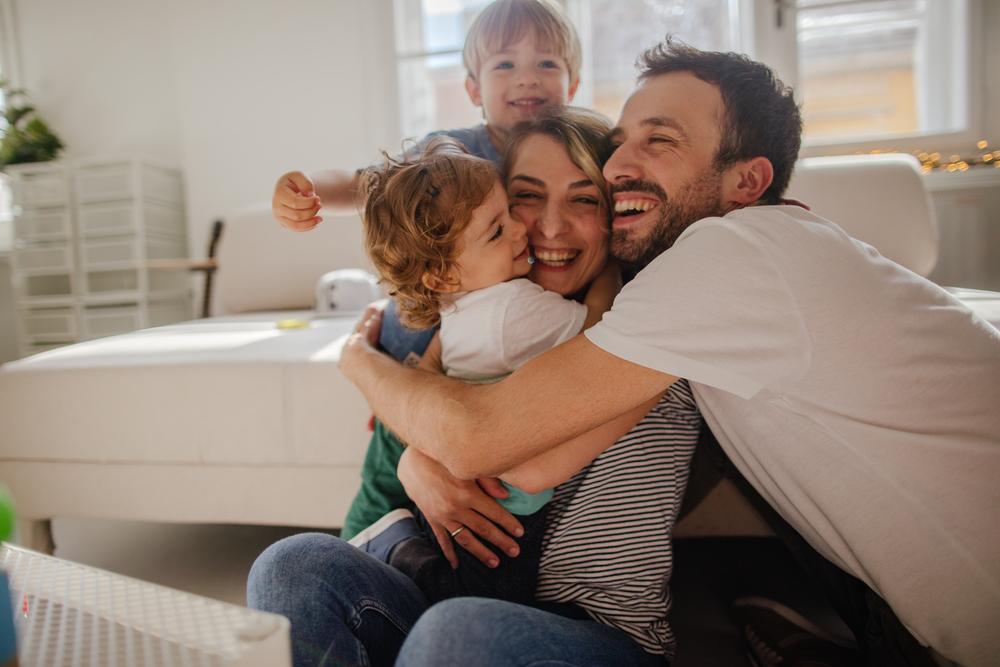 Família composta por pai, mãe e dois filhos de abraçando sentados no chão em frente ao sofá da sala