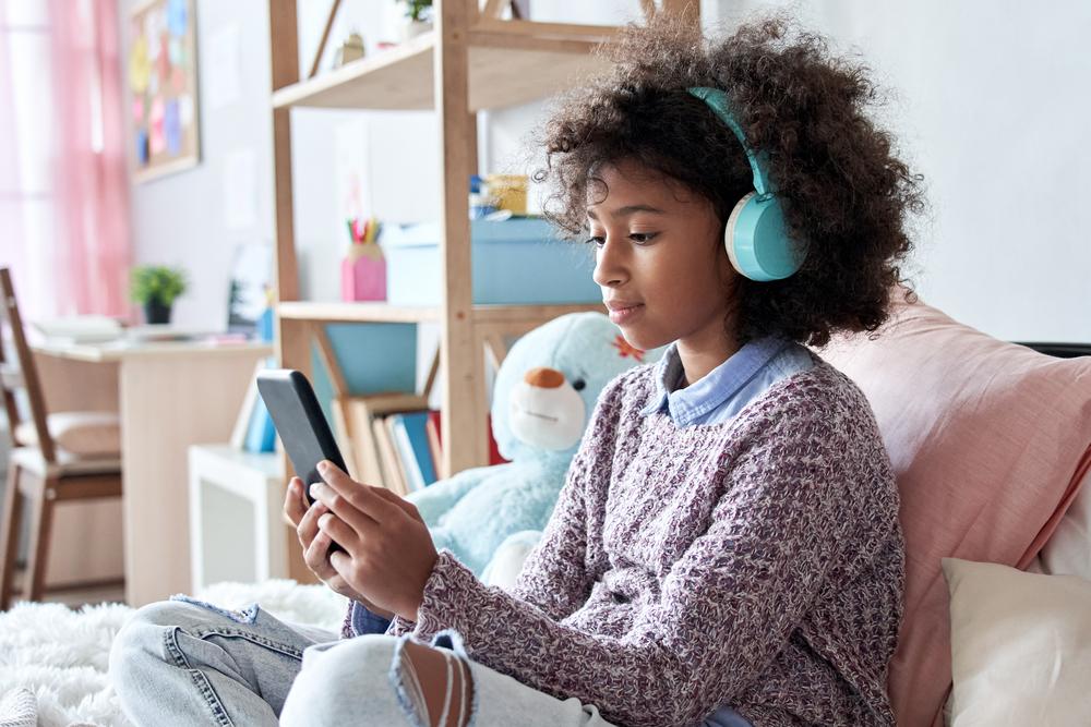 #ParaTodosVerem Menina negra pré-adolescente, com cabelo cacheado, solto, usando fones de ouvido azuis enquanto olha a tela do celular. Ela está sentada em uma cama com almofadas e veste blusão com calça jeans. O fundo da foto é um quarto de menina.