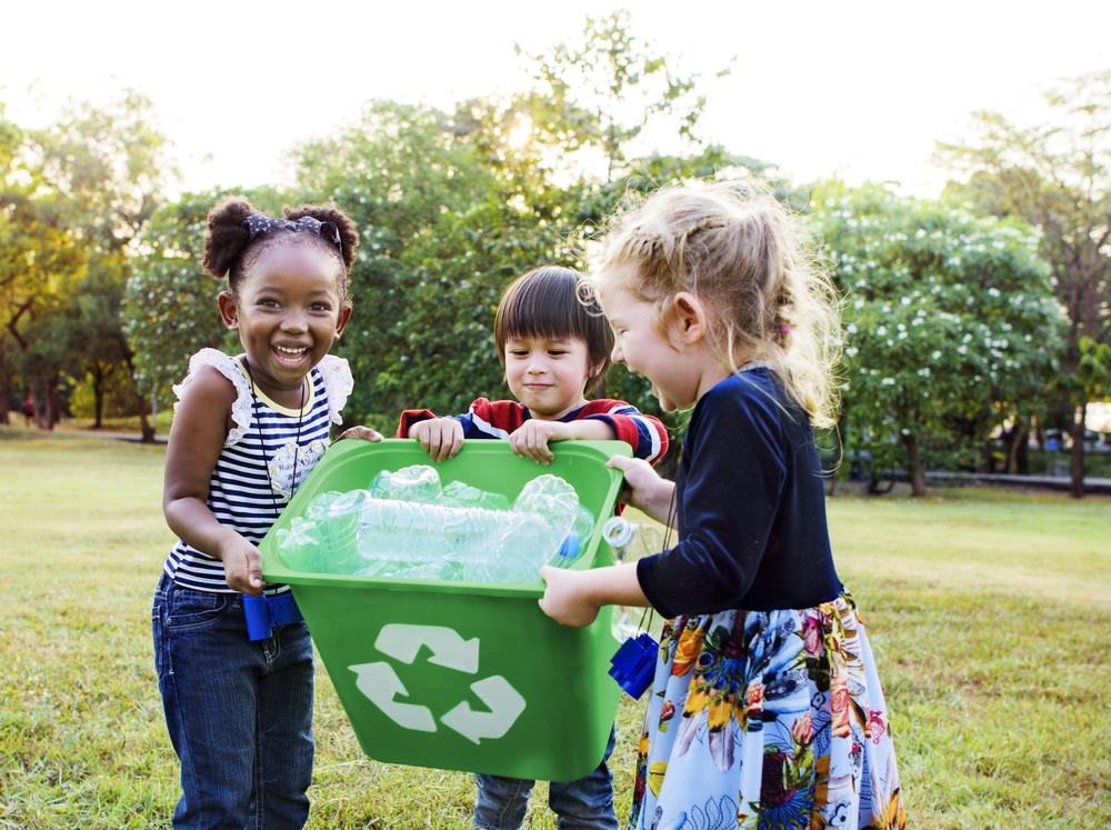 Imagem de três crianças, da esquerda para direita, uma menina, um menino e mais uma menina segurando um lixo com garrafas para reciclagem. Ao fundo, árvores e gramado.