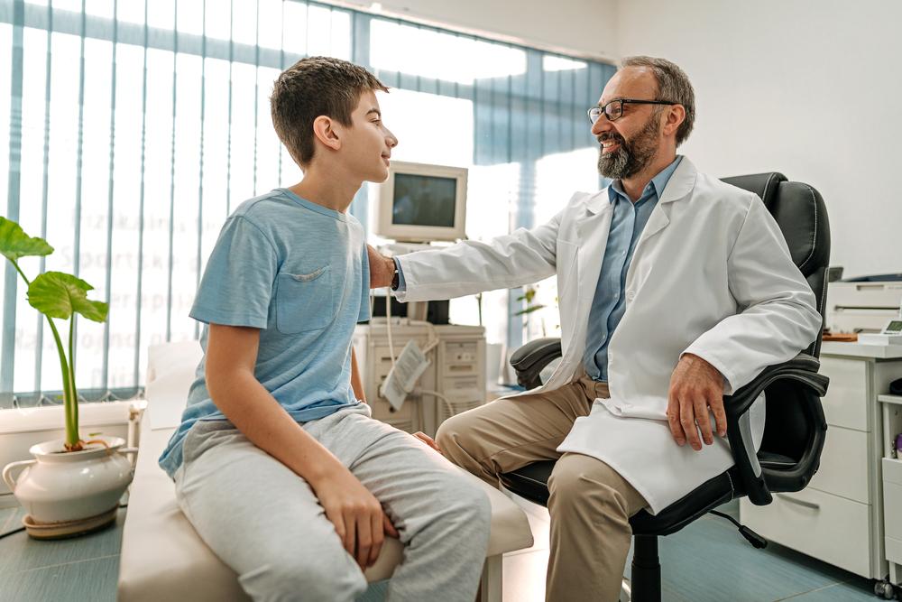 Imagem de um ambiente hospitalar em que um médico tranquiliza um paciente menino, ao colocar a mão no ombro dele. O médico está de jaleco branco, óculos e tem barba e o menino está vestindo uma camiseta azul