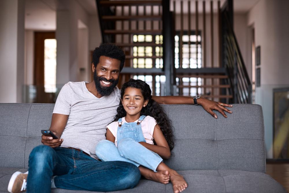 Em uma sala, um homem, vestindo uma camisa cinza, e uma menina, vestindo uma jardineira jeans e uma camisa branca, estão sentados em um sofá cinza. O homem está segurando um controle remoto, apontado para frente. O homem e a menina estão sorrindo. 