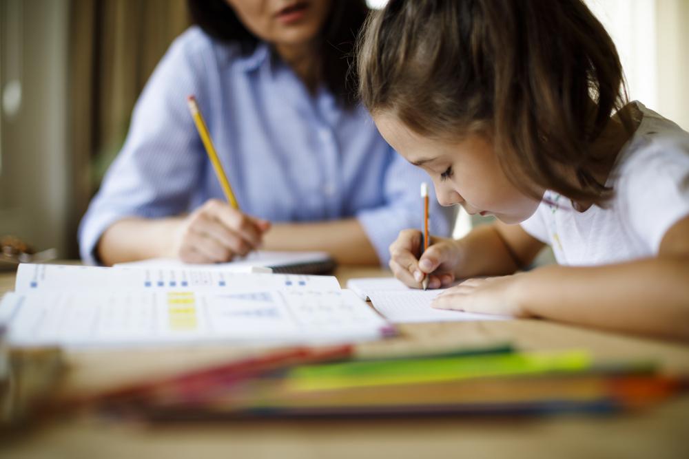 Sentados à mesa, estão uma mulher, que veste uma camisa azul, e uma menina, que veste uma camisa branca. A mulher e a menina estão escrevendo em cadernos, e a menina aparenta estar concentrada. Na mesa, estão alguns livros e canetas coloridas. 